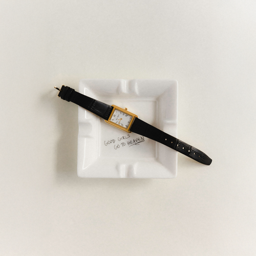 Watch with a black strap and gold case on a white dish with text that reads 'Good Girls Go To Heaven', against a light background